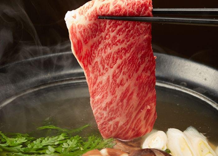 Marbled meat being dipped into a hot pot broth with mushrooms, green onions, and other veggies