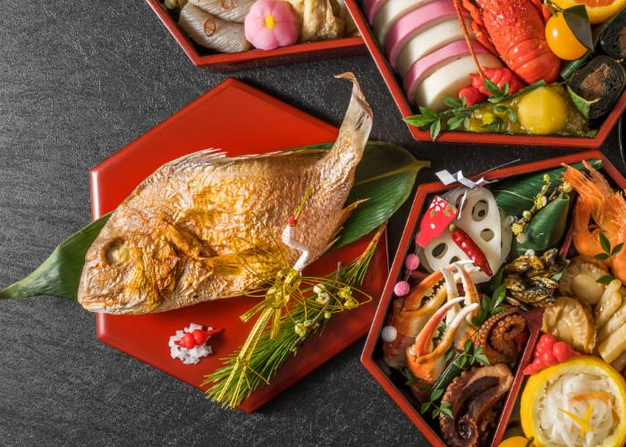 A whole cooked Tai sea bream on a red plate, next to boxes of carefully arranged osechi ryori, Japanese New Year foods