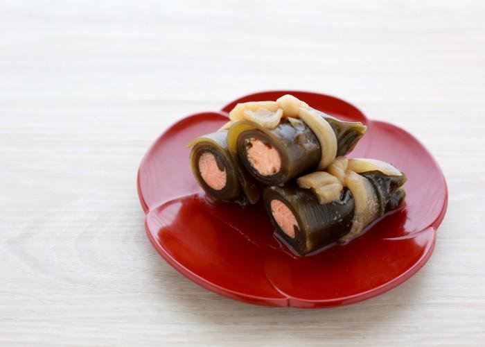 An osechi ryori dish called kombumaki, consisting of a wrapped piece of kombu kelp on a red plate