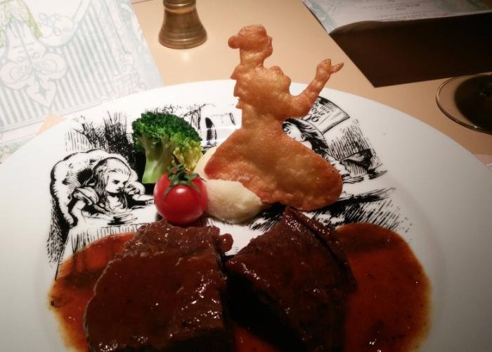 Slab of beef with with an Alice-shaped cracker on an Alice-printed plate at the Alice in Wonderland themed restaurant