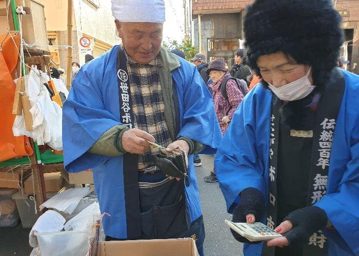 Old couple at their 50-year old stall with wooden shrine miniatures, the man is cashing in his wallet and the lady types on a calculator