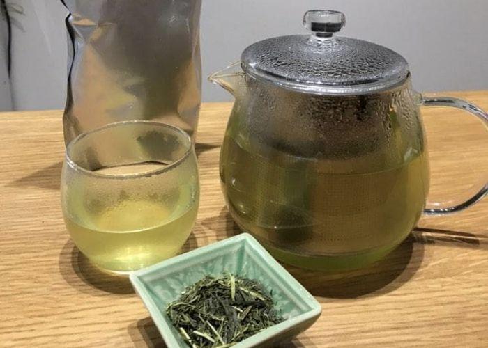 Glass teapot and cup with fresh brewed green tea, with some at display in a little ceramic four-edged plate.