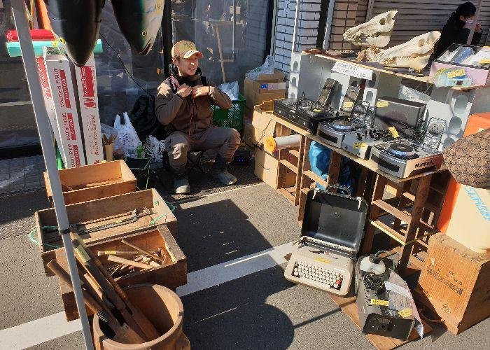 A man sits on a basket at his second hand stuff with farmer's tools on the left side and dj's equipment and a typing machine on the right side