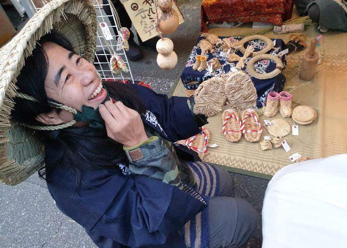 At the waraji (straw sandal) stall, the maker'S wife is showing me a pair of waraji and smiles broadly