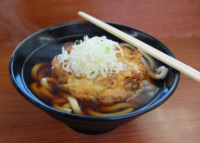A bowl of udon noodles in thin soy broth, topped with vegetable tempura and chopped spring onions.