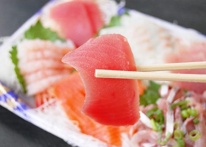 A piece of sashimi being held up to the camera, with a platter of pieces below.