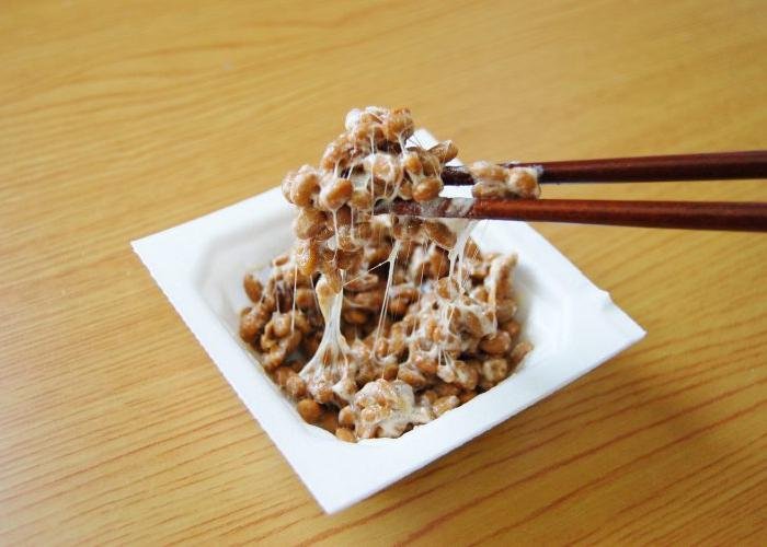A square bowl with a sticky and stringy mass of natto, being picked up with chopsticks.