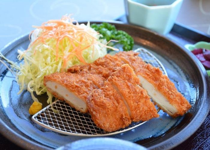 A katsu cutlet resting on a steel draining grill, with a pile of shredded cabbage and carrot on the side.
