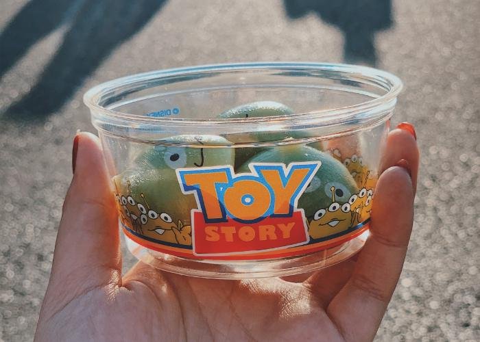Three Toy Story alien mochi is displayed inside a Toy Story themed cup. The cup is being held by a hand model. 