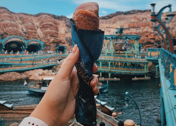 Freshly made turkey leg is wrapped with blue wrapping and held in the center of photo. In the background is Port of Discovery at Tokyo Disney Sea.