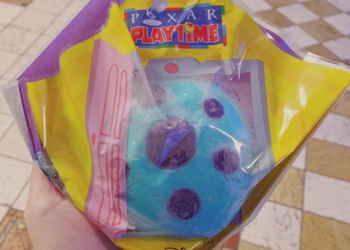 A blue bun with purple polka dots on it is covered with a Pixar playtime themed wrapping. The wrapping is clear in the middle to display the bun. 