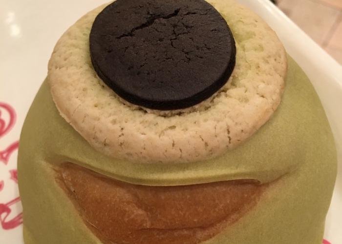 The pictured displayed shows Mike Wazowski Melon Pan up close. The main exterior is green and his smile is a light brown color. On the top is a round, white fluffy circular piece of bread and a chocolate cookie top to form the eye.