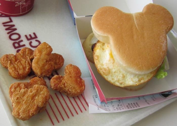 On the left is five crunchy chicken nuggets set on a tray. Next to it is a Mickey Mouse shaped burger held in a paper container. The burger has egg and salad sticking out on the side.