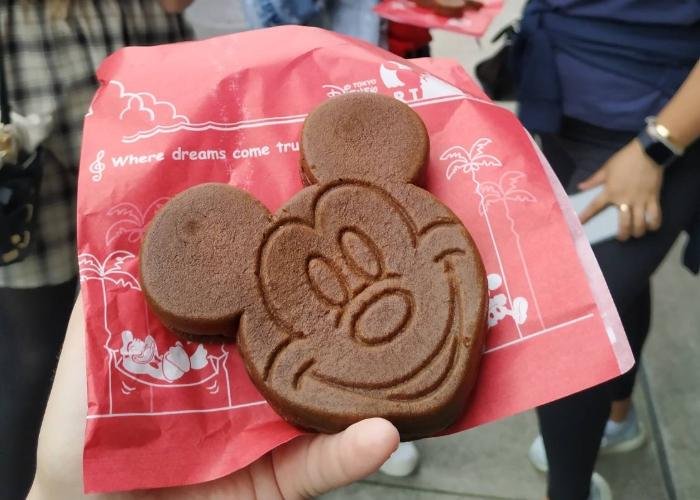 A brown, Mickey Mouse shape castella is placed on top of red parchment packaging. Mickey Mouse's face is engraved into the castella. On the red packaging towards the top, it says "where dreams come true".