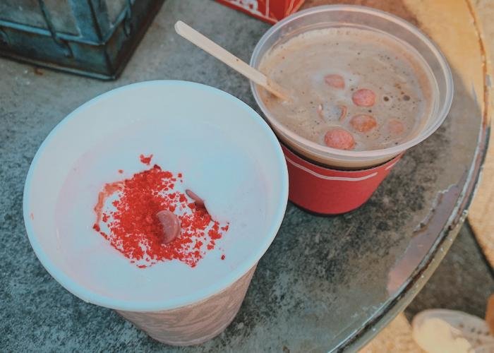 In this picture there are two drink. On the left is Raspberry Milk with with foam on top. Red sprinkles and pink chocolate chips are scattered on top of the foam. The right shows another drink, brown colored with a stir stick inside. 