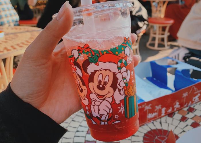 Inside a Mickey Mouse Christmas themed cup is red sparkling drink with boba balls setteled on the bottom. One the cup is Mickey and Minnie Mouse in Santa Claus outfits. There are gift boxes and snowflakes surrounding them.