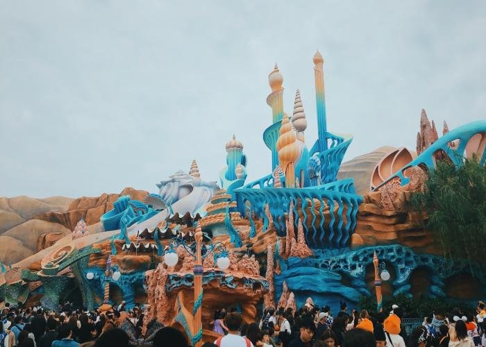 Ariel's castle is consisted of coral shaped structures with flow to represent the ocean. The color scheme is blue, white, and orange. There are people outside waiting to go into the castle. 
