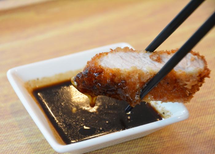 Piece of breaded tonkatsu meat being dipped into dish of brown sauce