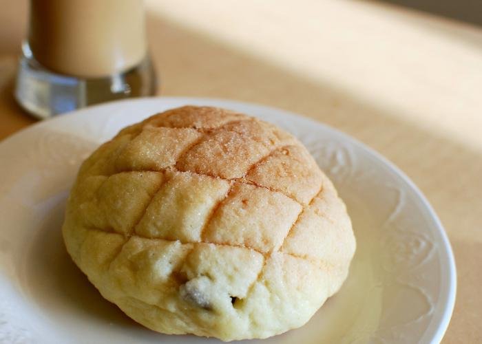 A fresh melon pan bun is placed on top of a white plate. There are checkered shaped grooves on top. In the background, there is a glass of coffee towards the top left side.