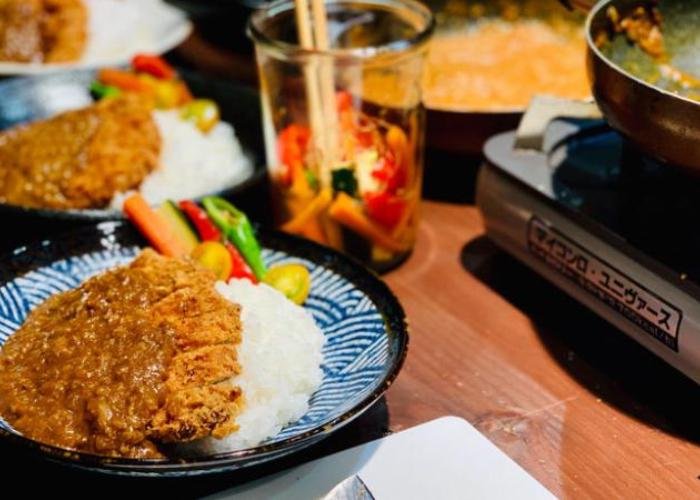 Plates of katsu curry are laid out in a row alongside a hot plate and metal cup of pickled vegetables. 