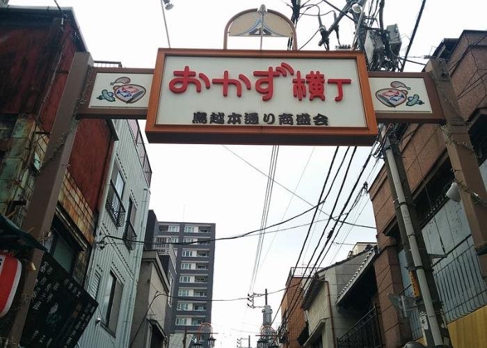 Okazu Yokocho, a drinking alley in Tokyo during the daytime with a large sign reading "Okazu Yokocho" in Japanese