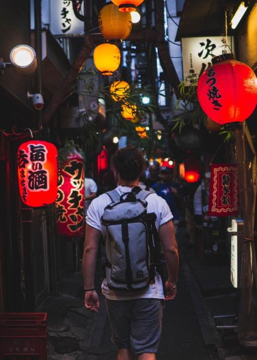 A man wearing a backpack walks through Omoide Yokocho drinking alley in Shinjuku at night with lanterns glowing