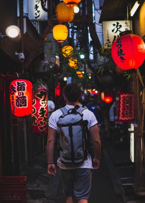 A man wearing a backpack walks through Omoide Yokocho drinking alley in Shinjuku at night with lanterns glowing