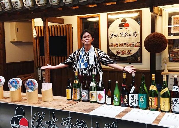 The barman of Fushimi Sakagura Koji stands behind the counter with his arms open, and 15 bottles in a row in front of him.