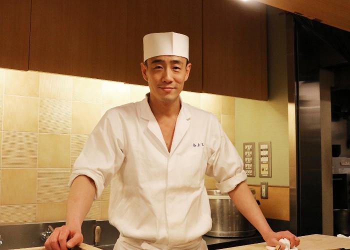 A young Japanese sushi chef in his white uniform leans on the wooden counter