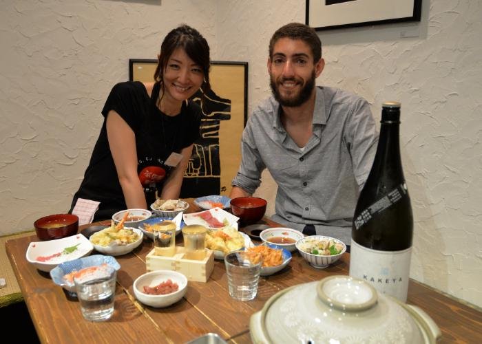 A man and a woman sitting at a table with a spread of Japanese food smile at the camera 