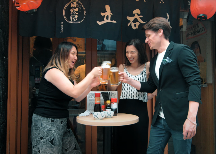 A man and two woman clink beer glasses outside a standing bar in Shibuya, with red hanging lanterns outside
