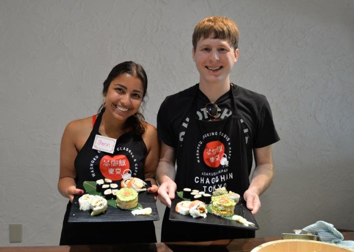 A man and a woman hold out trays of their sushi rolls and mille-feuille sushi