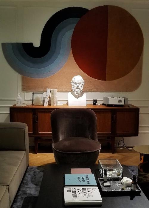 A couch sits to the left with a coffee table and books in front of it. To the back is a colorful wall hanging and the bust of a man