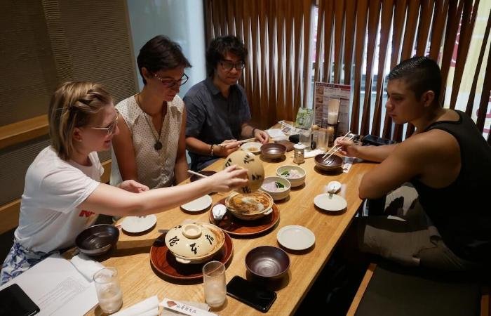 Travelers sat around table with dishes, one opening pot of miso nikomi udon